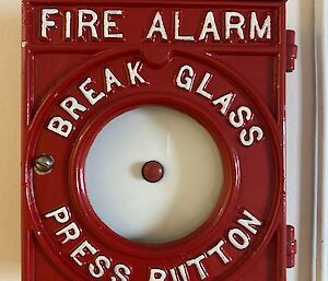 An antique fire alarm. The casing is painted red with white letters reading "Fire alarm - break glass - press button". The red button is behind a circular pane of glass