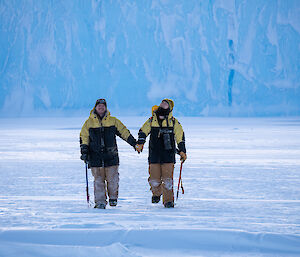 Two men walk across sea-ice holding hands, with iceberg in background