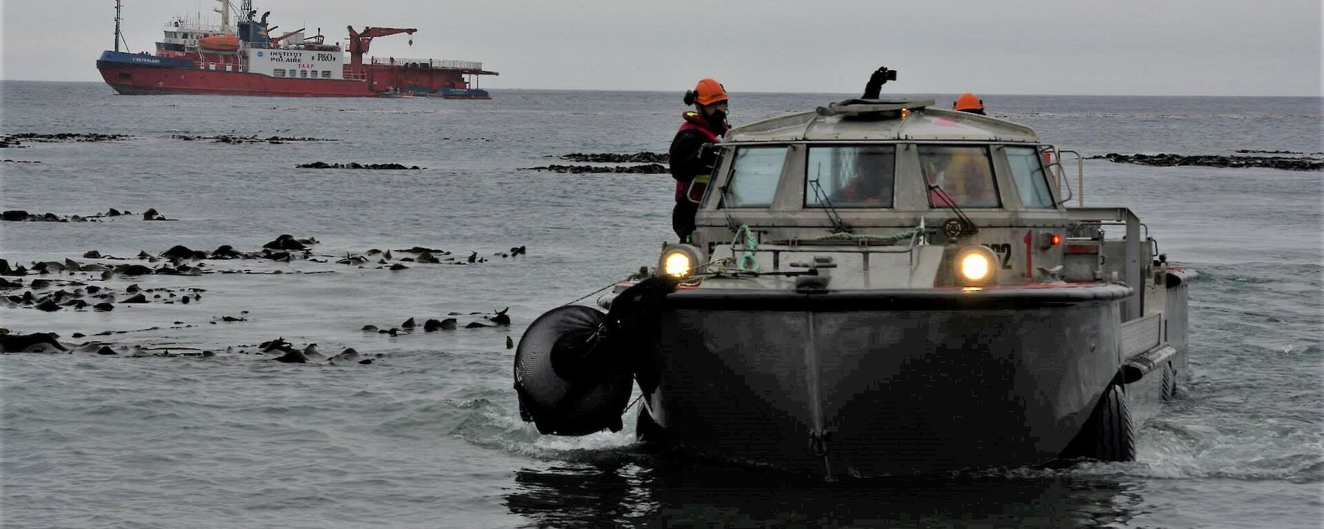 A silver barge comes ashore as a red and white ship sits offshore