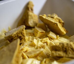 A close-up view of broken pieces of honeycomb confectionery. The light yellow chunks vary in size, have jagged edges and a porous texture