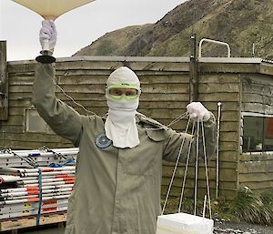 A man in a protective coat, fireproof mask and goggles holds up a large white weather balloon