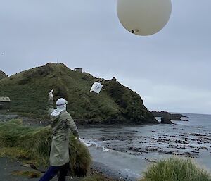 A man in protective clothing launches a large white weather balloon in to the air near the shore