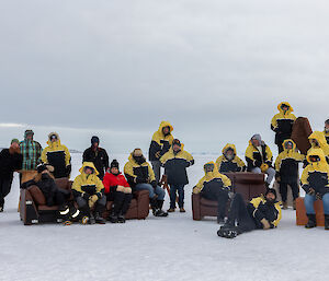 A group of people sitting and standing on furniture on the sea ice with yellow jackets on
