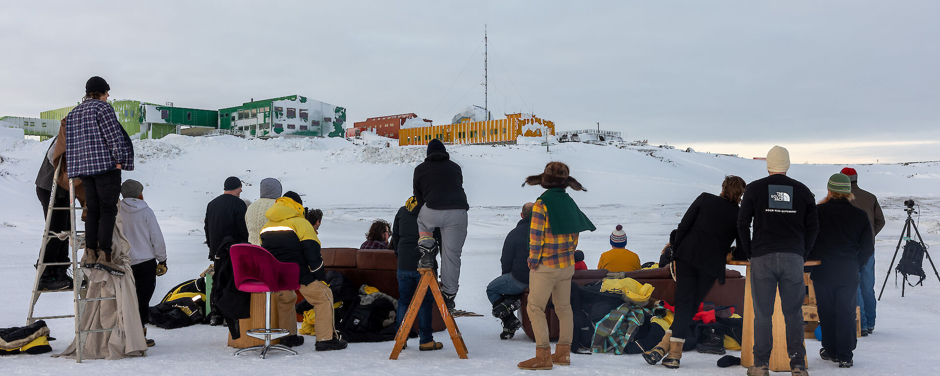 A group of people standing and sitting on furniture on the sea ice taken from behind