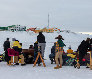 A group of people standing and sitting on furniture on the sea ice taken from behind