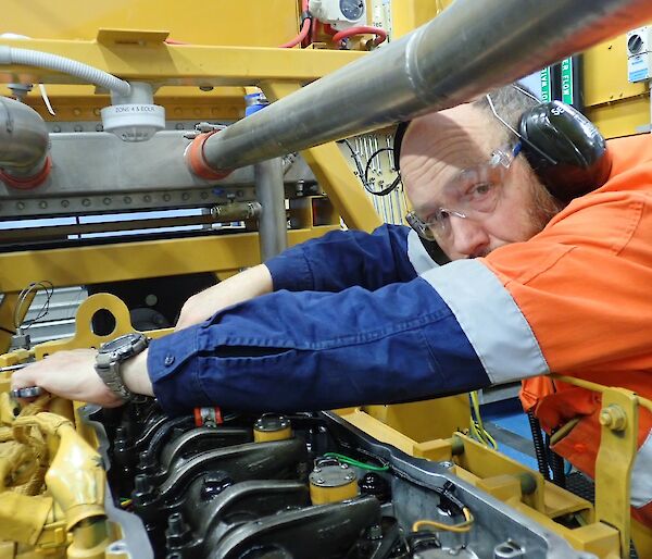 Man in high vis and wearing protective glasses and ear protection leans over a large engine and conducts adjustment.