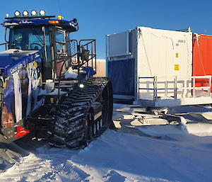 A tractor hitched to a row of large cargo containers mounted on sleds, ready to pull them across the snow