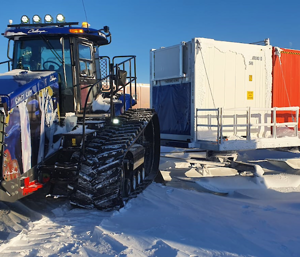 A tractor hitched to a row of large cargo containers mounted on sleds, ready to pull them across the snow