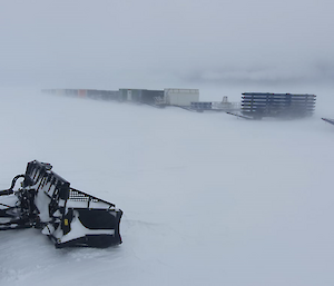 Cargo containers mounted on sleds, arranged in a row on snowy ground. The row of container fades into a white, blurry distance, suggesting that the air is full of snow blown in a very strong wind. In the foreground, a snow groomer is smoothing a path through the snow.