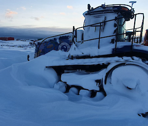 A tractor parked on snowy ground. Its tracks and wheels are caked and half-buried in massive deposits of snow. A coating of blown snow also covers its windows.