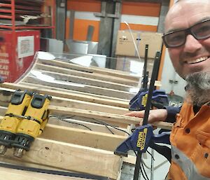 A selfie taken by a man in an orange work shirt and glasses, showing him standing at a workbench, working on a curved pane of transparent material.