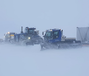 Two snow groomers, a tractor and a cube-shaped cabin on a sled in a snowy location. They are in 'white-out' conditions, where so much snow is being blown by the wind that the ground is indistinguishable from the sky.