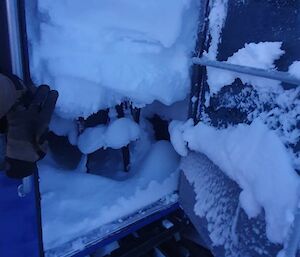 A view through the open door of a cabin, showing large deposits of blown snow built up inside, and even small icicles hanging from the ceiling.