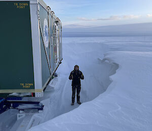 A man is standing in a wind scour: a mini-canyon carved by strong winds into the snow around a caravan cabin mounted on sleds. The scour is about 30 centimetres deeper than the man is tall.