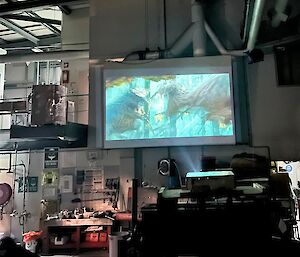 View from seat with feet up and dinosaurs battling on the large cinema screen, all in the workshop environment