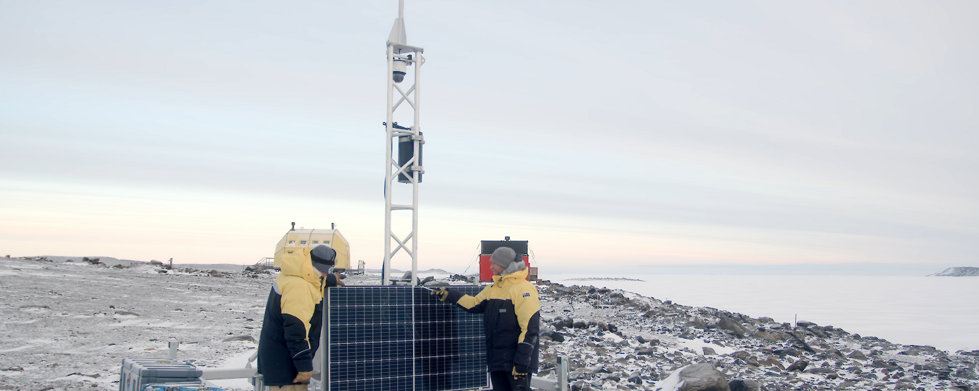 Two men in yellow jackets stand next to solar panels connected to a metal tower