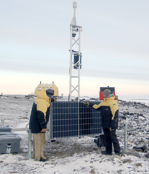 Two men in yellow jackets stand next to solar panels connected to a metal tower