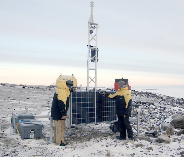 Two men in yellow jackets stand next to solar panels connected to a metal tower