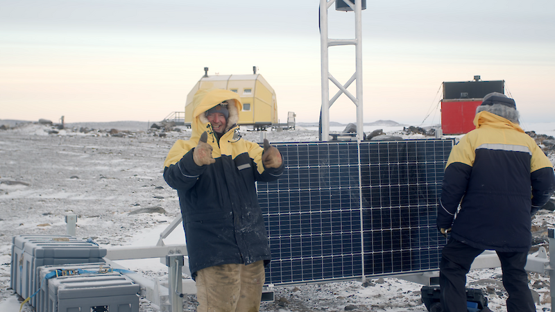 Two men in yellow jackets smile stand next to a solar panel attached to a small metal tower. One man is laughing and giving the thumbs up