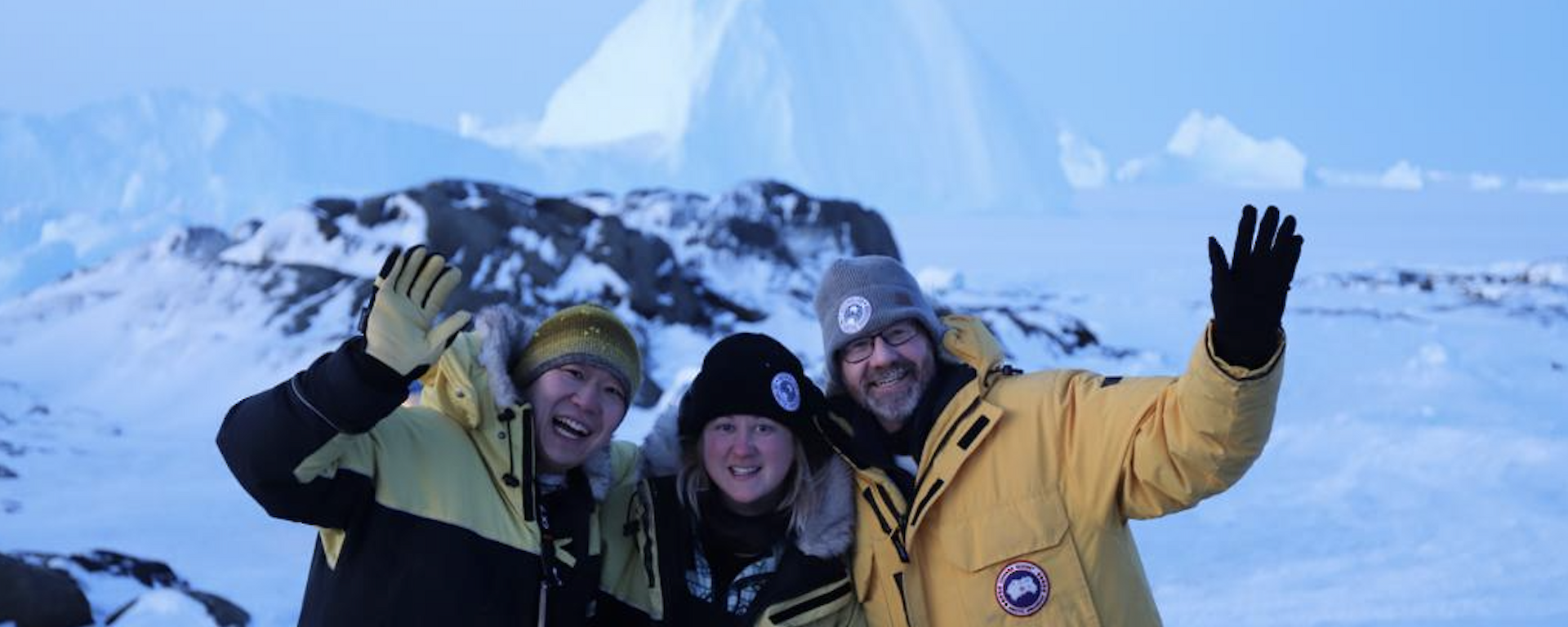 Three people in cold weather gear, standing in front of a big ice berg at Macey
