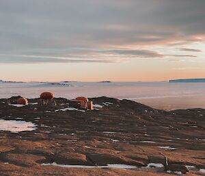 In foreground rocky uneven ground, in the middle distance the field camp made up of two googies and a melon and in the distance sea ice to the horizen