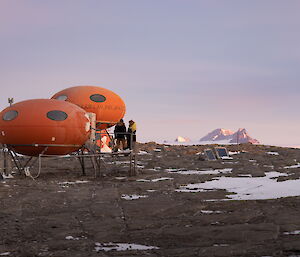 In the foreground two red googies with two expeditioners standing on the verandah of the closest, and two mountain peaks rise up on the distant horizen