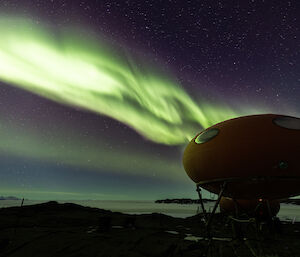 In the foreground a googie hut at night, above a very bright green aurora emits a green glow across the whole scene