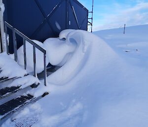 Steel steps leading up to a shed in a snowy location. Beside the steps and against the wall of the shed, snow has been blown and piled by strong winds into the shape of an ocean wave