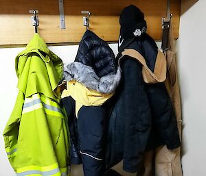 A corner of a cold porch, with three thick winter jackets and a pair of overalls hung up on hooks, a few pairs of boots beneath them, and a pair of thick gloves, a water bottle and a 2-way radio lying on a bench