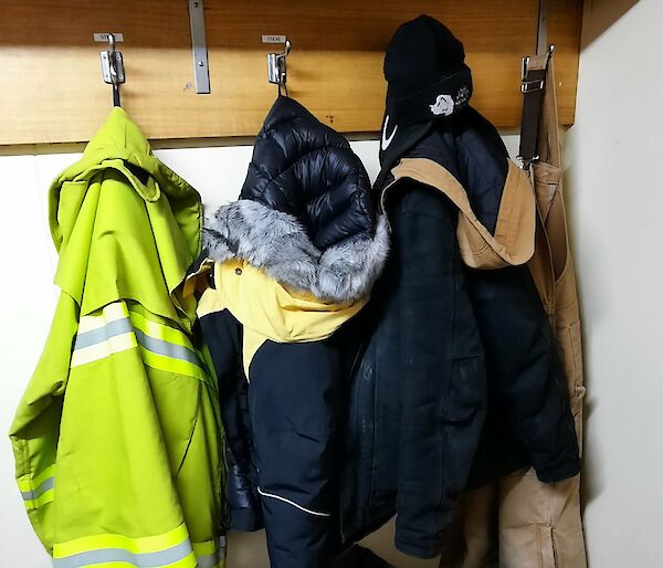 A corner of a cold porch, with three thick winter jackets and a pair of overalls hung up on hooks, a few pairs of boots beneath them, and a pair of thick gloves, a water bottle and a 2-way radio lying on a bench