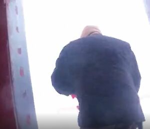 A view from behind of a man in thick, protective winter clothing going out through a door. "White-out" weather conditions obscure the view outside - nothing can be distinguished in the uniform, blank whiteness