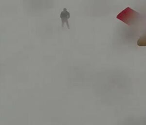 A photo taken in a blizzard. Nothing can be distinguished in the blank, greyish whiteness, except for a gloved hand reaching out with a smartphone, and the hazy silhouette of a person about 20 metres ahead
