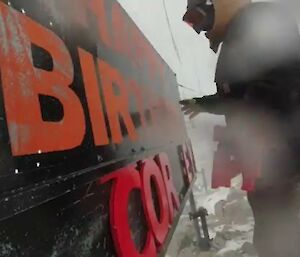 A man in thick, protective winter clothing placing letters up on a letter board. The photo is slightly hazy due to the amount of blowing snow when it was taken