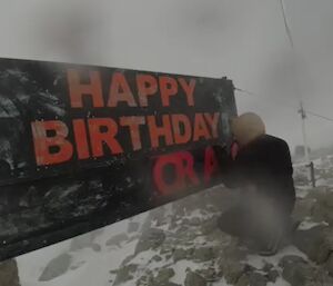 A man in thick, protective winter clothing, crouching beside a letter board to place the letters of someone's name. Above the slot for the letters are the words "HAPPY BIRTHDAY" in bright orange. The photo is blurred in places due to the amount of blowing snow when it was taken