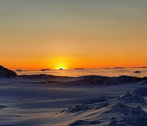A sunset scene in Antarctica. Snow in the foreground has been shaped into low dunes and ridges by strong winds. In the background, the ice-covered ocean is lit palely by the sun setting in an amber sky