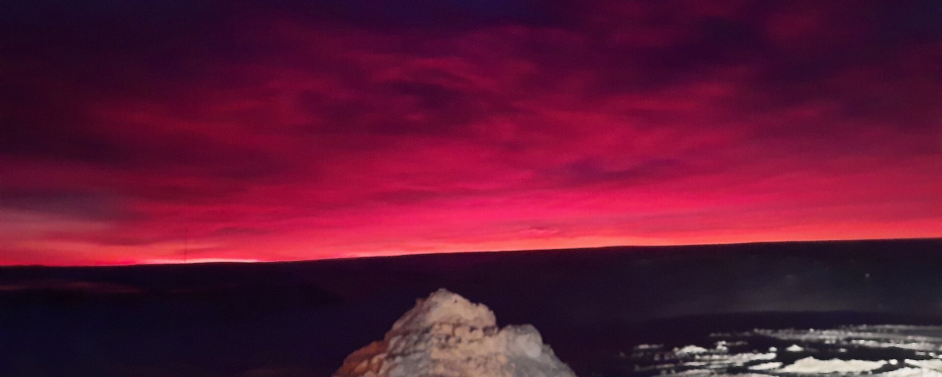 A sunrise scene in Antarctica. In the foreground, a pile of snow is lit up by station lighting. In the background, the cloudy sky is magenta and purple with dawn colours