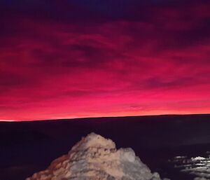 A sunrise scene in Antarctica. In the foreground, a pile of snow is lit up by station lighting. In the background, the cloudy sky is magenta and purple with dawn colours