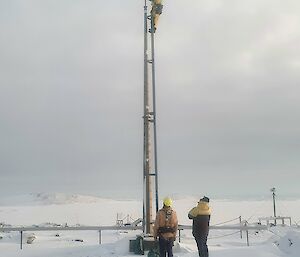 A man on a mast while 2 others look on for safety amidst the snow