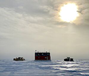 A small, rectangular building mounted on sleds, being towed by a tractor across an icy plain. Another tractor is hitched to the rear of the building. The sky is greyish-white with layers of high cloud, behind which the sun shines brightly
