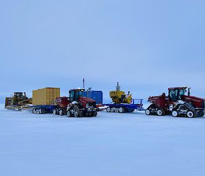 Two red tractors driving across a flat, snowy plain beneath a cloudy sky. They are both towing flatbeds loaded with blue and yellow cargo containers and large machinery items.