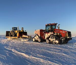 A red tractor driving across a flat, snowy plain beneath a clear, blue sky. It is towing a sled assembly on which sits a smaller, yellow tractor