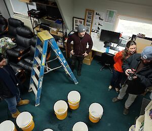 A group of people are surrounded by white buckets on the floor and a ladder