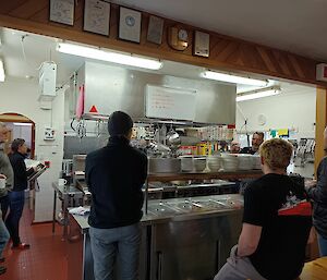 A group of people stand around watching others in a kitchen