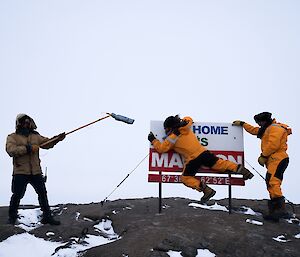Man is blown against sign reading 'its home its Mawson' while 2nd man stands at left holding boom and 3rd man stands at right looking on