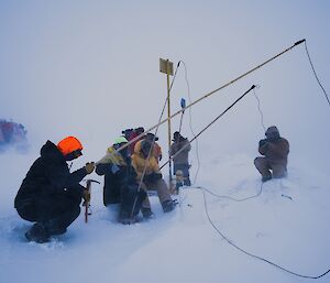 Group of three in blizzard, sit on buckets and hold fishing lines are over looked by three production crew members