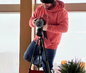 Man stands on chair precariously holding camera on tripod filming food on a plate on table from above