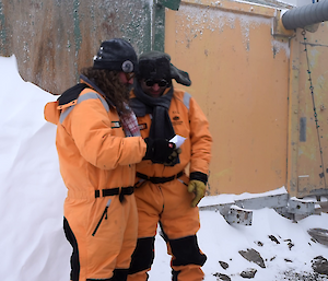 Two men stand in yellow freezer suits outside old building looking at a piece of paper