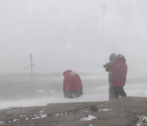 Man in red jacket kneels in snow with back to camera, two production crew look on, blizz blowing snow across the scene