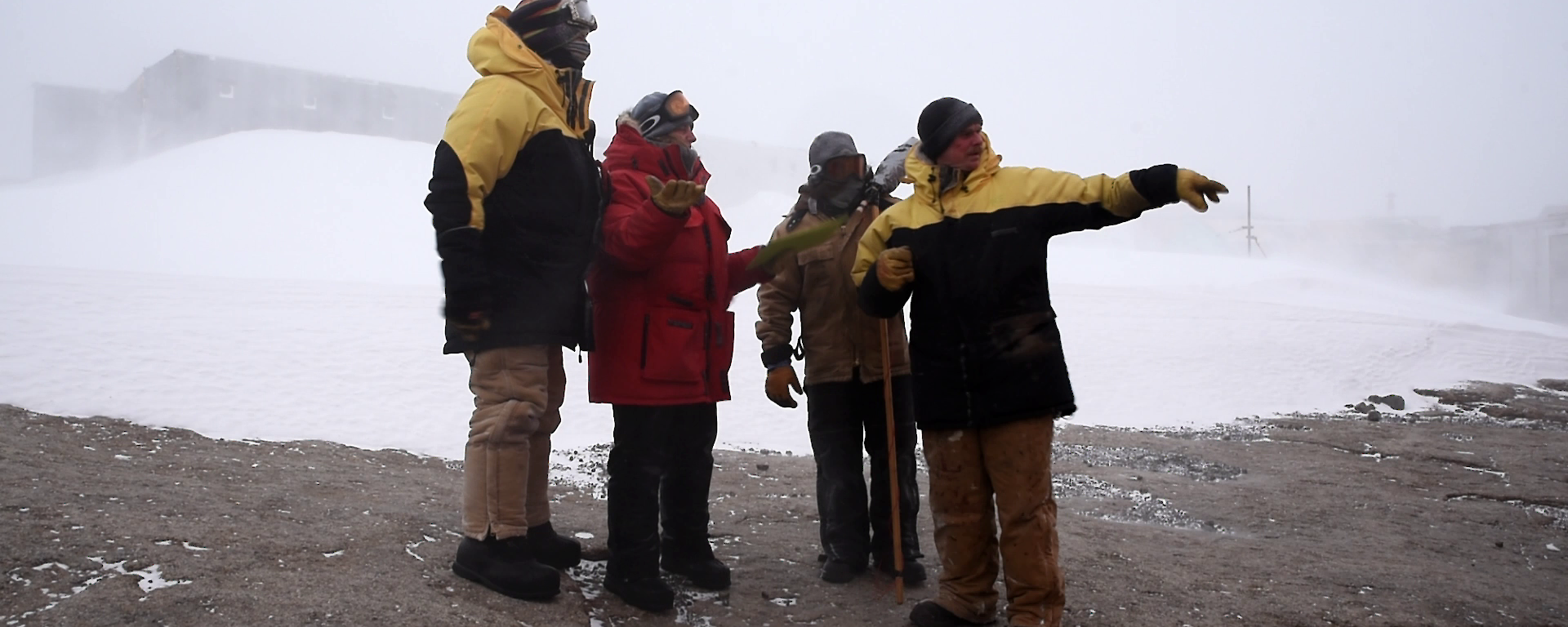 Four people standing in a huddle talking, foreground rock and background ice and buildings obstructed by blizzard