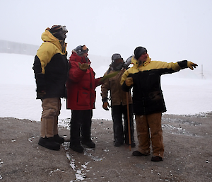 Four people standing in a huddle talking, foreground rock and background ice and buildings obstructed by blizzard